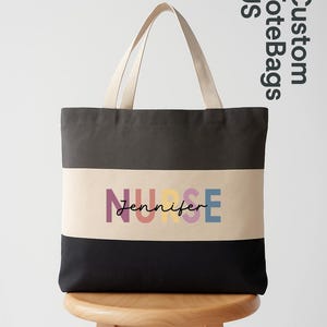 May include: A black, grey, and beige tote bag with the text "NURSE Jennifer" in colorful letters. The bag has beige handles and is sitting on a wooden stool. The words "Custom Tote Bags US" are in the upper right corner.