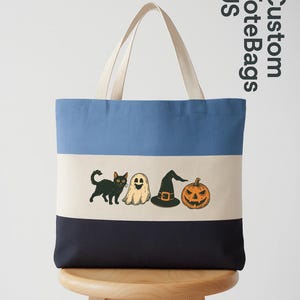 May include: A canvas tote bag with a blue, off-white, and navy color block design. The bag features Halloween-themed illustrations of a black cat, ghost, witch hat, and jack-o'-lantern. The bag has off-white handles and the text "Custom Tote Bags US".