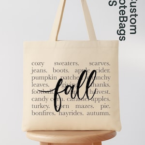 May include: Cream-colored tote bag with the word "fall" in a cursive font. The bag features a list of fall-themed words like "cozy," "sweaters," and "apple cider." The bag is on a wooden stool.