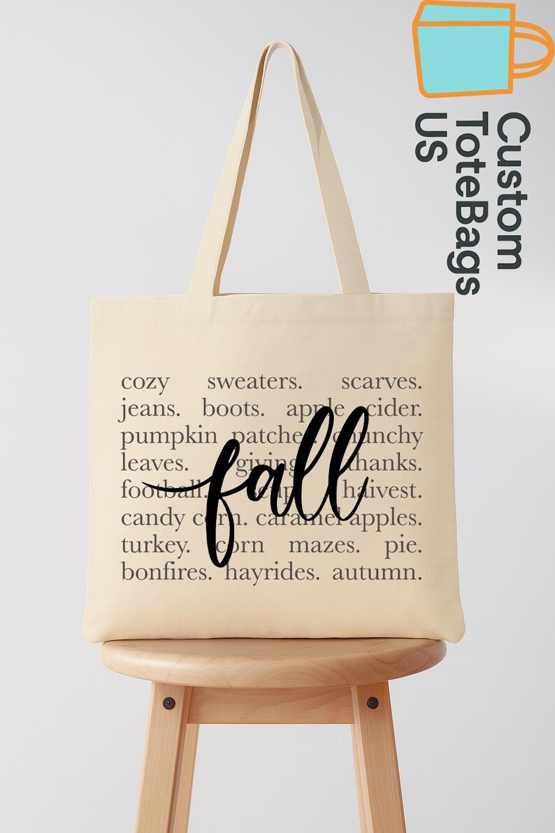 May include: Cream-colored tote bag with the word "fall" in a cursive font. The bag features a list of fall-themed words like "cozy," "sweaters," and "apple cider." The bag is on a wooden stool.