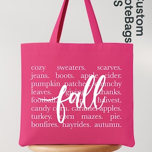 May include: A bright pink tote bag with white text listing fall-themed words like "cozy," "sweaters," and "autumn." The word "fall" is written in a cursive font across the center. The bag has long handles and the words "Custom Tote Bags US" in the upper right corner.