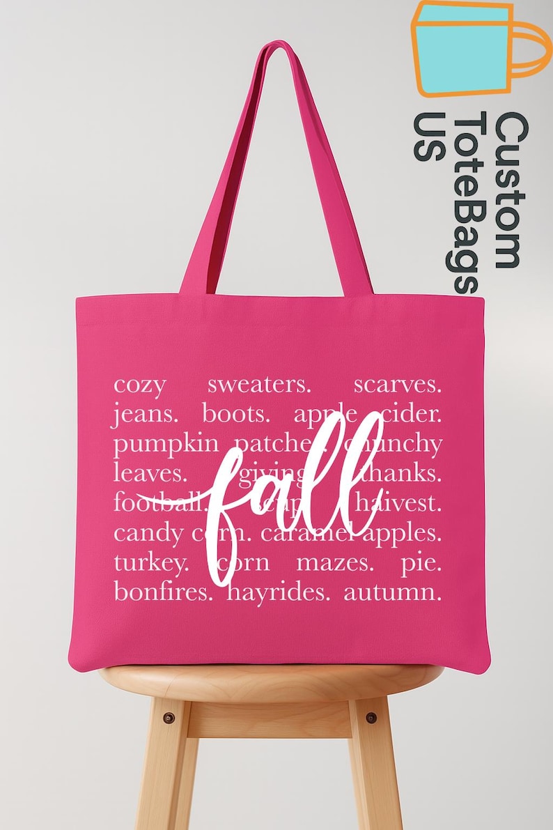 May include: A bright pink tote bag with white text listing fall-themed words like "cozy," "sweaters," and "autumn." The word "fall" is written in a cursive font across the center. The bag has long handles and the words "Custom Tote Bags US" in the upper right corner.