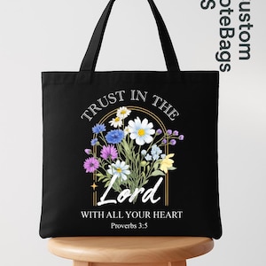 May include: A black tote bag featuring a floral graphic with white, blue, purple, and yellow flowers. The text reads: TRUST IN THE LORD WITH ALL YOUR HEART Proverbs 3:5. The design is centered on the front of the bag with two black shoulder straps.