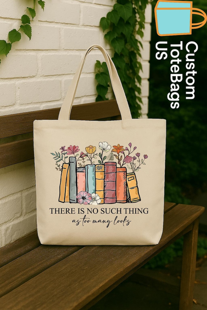 May include: Cream-colored tote bag featuring a colorful illustration of books with flowers on top. The text "THERE IS NO SUCH THING as too many books" is printed below the books. The bag has two handles and is sitting on a wooden bench.