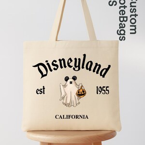 May include: Beige tote bag with black text that reads "Disneyland est 1955 CALIFORNIA". A cartoon ghost with Mickey Mouse ears and a jack-o'-lantern is in the center. The bag has long beige handles.