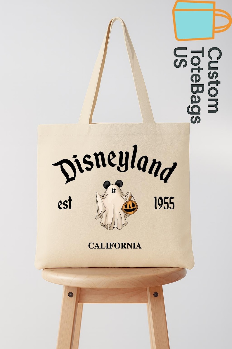 May include: Beige tote bag with black text that reads "Disneyland est 1955 CALIFORNIA". A cartoon ghost with Mickey Mouse ears and a jack-o'-lantern is in the center. The bag has long beige handles.