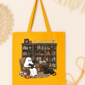 Puede incluir: Bolso tote amarillo mostaza con una ilustraci&oacute;n de un fantasma leyendo en una biblioteca. El dise&ntilde;o incluye un gato negro, calabazas y una estanter&iacute;a con libros, velas y una calavera. El bolso tiene una forma rectangular simple.