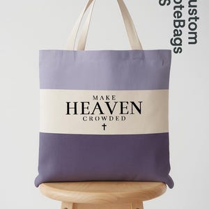 May include: A canvas tote bag with a lavender and purple color block design. The bag features the text "MAKE HEAVEN CROWDED" in black, with a cross symbol below. The handles are a cream color.