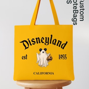 May include: A yellow tote bag with black lettering that says "Disneyland est 1955 CALIFORNIA". The bag features a cartoon ghost holding a jack-o'-lantern with Mickey Mouse ears. The bag has yellow handles and is sitting on a wooden stool.