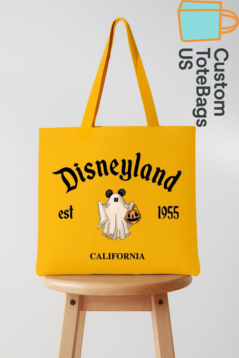 May include: A yellow tote bag with black lettering that says "Disneyland est 1955 CALIFORNIA". The bag features a cartoon ghost holding a jack-o'-lantern with Mickey Mouse ears. The bag has yellow handles and is sitting on a wooden stool.