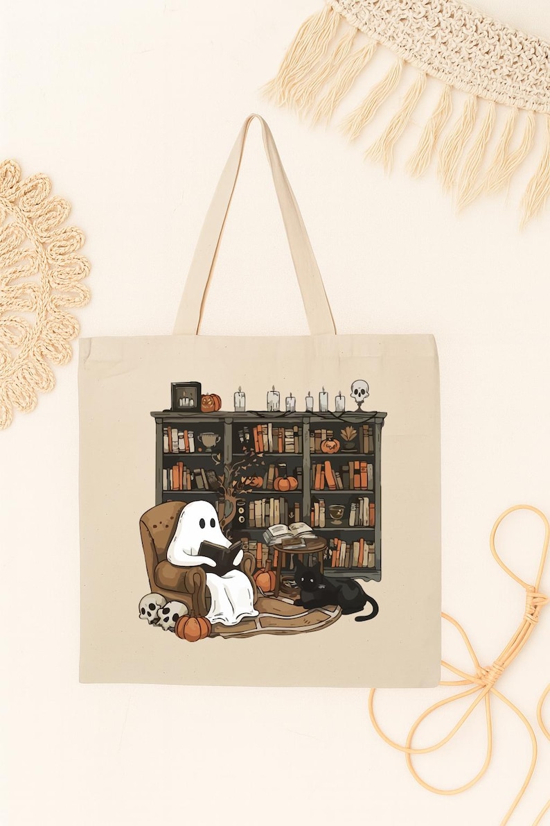 Puede incluir: Bolso tote beige con una ilustraci&oacute;n de Halloween. Un fantasma lee en una silla frente a una estanter&iacute;a, con un gato negro, calabazas y calaveras. El bolso tiene un fondo y asas color crema.