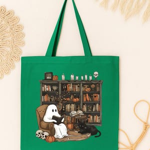 Puede incluir: Bolso tote verde con una ilustraci&oacute;n caprichosa de un fantasma leyendo en una biblioteca. La escena incluye un gato negro, calabazas, velas y una estanter&iacute;a llena de libros. Un dise&ntilde;o divertido y espeluznante para los amantes de los libros.