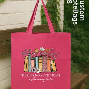 May include: A pink tote bag featuring a colorful illustration of books with flowers growing out of them. The text "THERE IS NO SUCH THING as too many books" is below the illustration. The bag has pink handles.