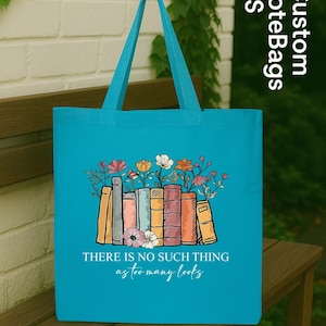 May include: A turquoise tote bag with a design of books and flowers. The text "THERE IS NO SUCH THING as too many books" is printed on the bag. The bag has turquoise handles and is on a wooden bench.
