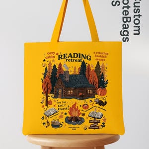 May include: Mustard yellow tote bag featuring a cabin scene with the words "Reading Retreat." The design includes a cabin, trees, a campfire, and the text "For the Cozy Readers." The bag has yellow handles and the text "Custom Tote Bags US."