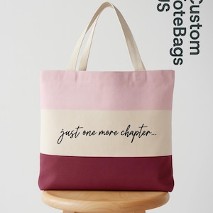 May include: A multi-colored tote bag with a pink, cream, and burgundy striped design. The bag features the text "just one more chapter..." in a handwritten font. The bag has cream-colored handles and is sitting on a wooden stool.
