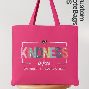 May include: A bright pink tote bag with the words "KINDNESS is free" in colorful letters. The bag has long handles and a leopard print heart graphic. The phrase "SPRINKLE IT EVERYWHERE" is printed below.