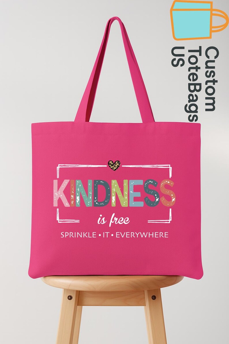 May include: A bright pink tote bag with the words "KINDNESS is free" in colorful letters. The bag has long handles and a leopard print heart graphic. The phrase "SPRINKLE IT EVERYWHERE" is printed below.