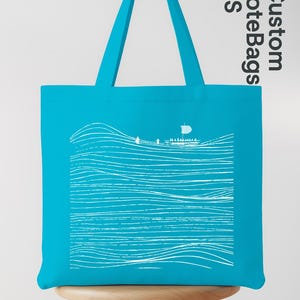 Puede incluir: Bolso tote turquesa con un dise&ntilde;o de olas y barco en blanco. El bolso tiene forma rectangular con asas a juego. El texto "Custom Tote Bags US" est&aacute; impreso en el lateral. El bolso est&aacute; sobre un taburete de madera.