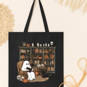 Puede incluir: Bolso tote negro con una ilustraci&oacute;n de Halloween. Representa un fantasma leyendo en una biblioteca, rodeado de calabazas, velas, una calavera y un gato negro. El bolso tiene asas negras.