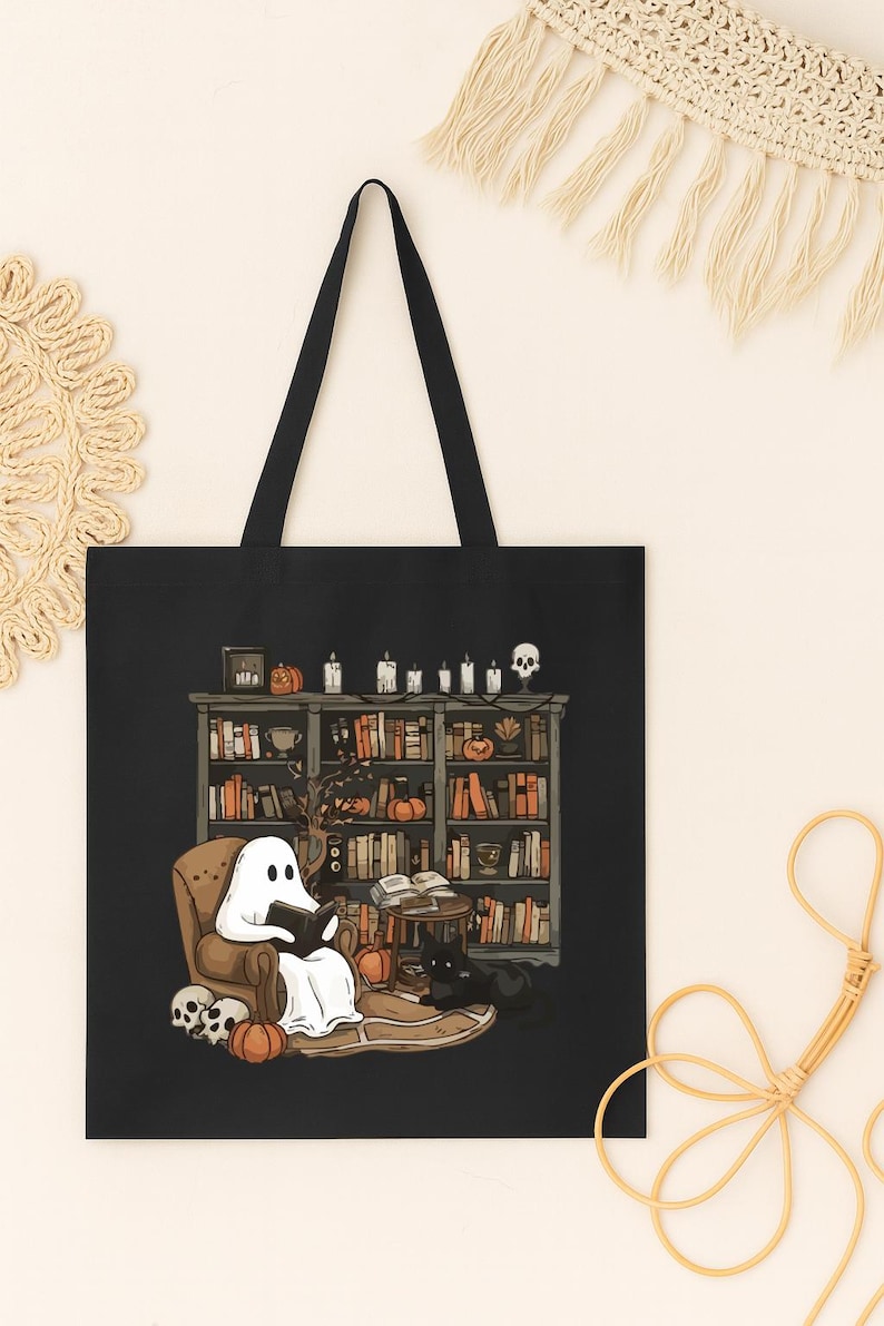 Puede incluir: Bolso tote negro con una ilustraci&oacute;n de Halloween. Representa un fantasma leyendo en una biblioteca, rodeado de calabazas, velas, una calavera y un gato negro. El bolso tiene asas negras.