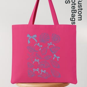 K&ouml;nnte beinhalten: Eine leuchtend pinkfarbene Tragetasche mit blauen Muschel- und Schleifenillustrationen. Die Tasche hat lange Henkel und der Text "Custom Tote Bags US" ist auf der Seite aufgedruckt. Die Tasche steht auf einem Holzschemel.