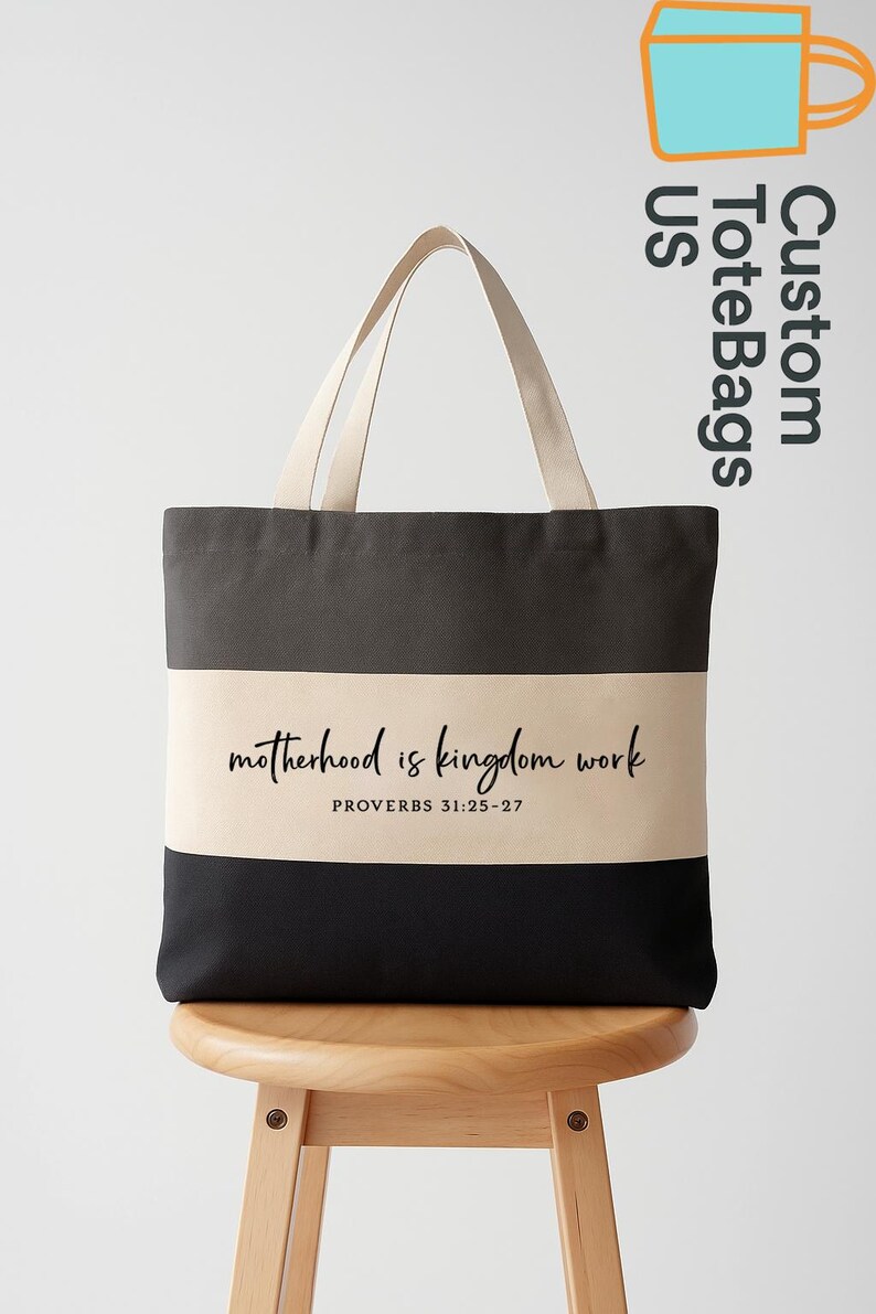 May include: A black, gray, and beige tote bag with the text "motherhood is kingdom work" and "PROVERBS 31:25-27". The bag has beige handles and is sitting on a wooden stool.