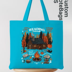 May include: A turquoise tote bag featuring a cabin scene with the words "READING retreat." The design includes a cabin, trees, a campfire, and the text "For the cozy readers." The bag has a handle.