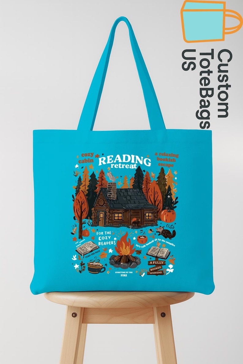 May include: A turquoise tote bag featuring a cabin scene with the words "READING retreat." The design includes a cabin, trees, a campfire, and the text "For the cozy readers." The bag has a handle.