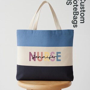 May include: A striped tote bag with a blue top, off-white center, and navy bottom. The word "NURSE" is printed in colorful letters above the name "Jennifer". The bag has off-white handles and is sitting on a wooden stool.