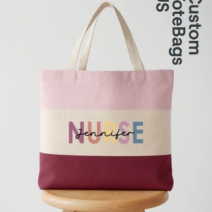 May include: A custom tote bag with a pink, cream, and burgundy color block design. The word "NURSE" is printed in colorful letters, with the name "Jennifer" below. The bag has cream-colored handles.