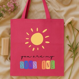 Peut inclure: Un sac fourre-tout rose vif avec un soleil jaune et l'inscription "you are my sunshine" en &eacute;criture noire. Le mot "sunshine" est en lettres color&eacute;es. Le sac a des poign&eacute;es roses assorties.