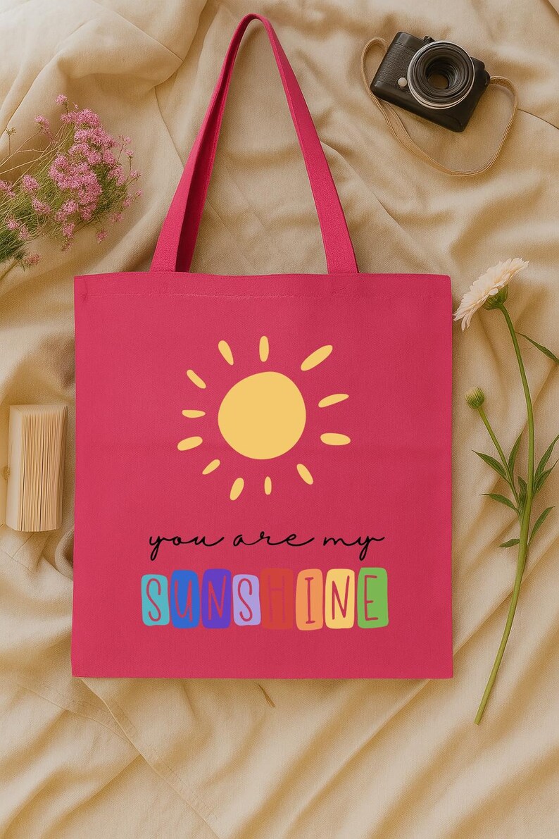 Peut inclure: Un sac fourre-tout rose vif avec un soleil jaune et l'inscription "you are my sunshine" en &eacute;criture noire. Le mot "sunshine" est en lettres color&eacute;es. Le sac a des poign&eacute;es roses assorties.