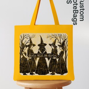 May include: A yellow tote bag with a graphic of five women in witch hats and dresses, with the text "Girls Will Be Girls." The bag has matching yellow handles and is displayed on a wooden stool. The bag is a vibrant yellow.
