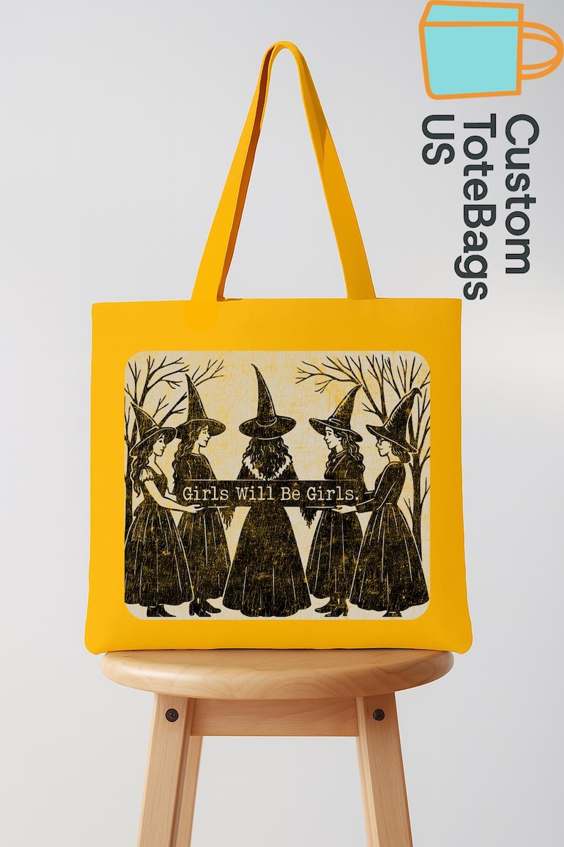 May include: A yellow tote bag with a graphic of five women in witch hats and dresses, with the text "Girls Will Be Girls." The bag has matching yellow handles and is displayed on a wooden stool. The bag is a vibrant yellow.
