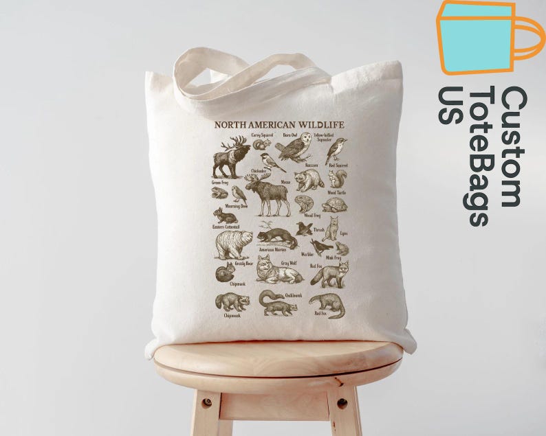May include: A natural-toned tote bag displaying a detailed illustration of North American wildlife. The design includes various animals such as deer, bears, and birds, with the text "NORTH AMERICAN WILDLIFE" at the top. The bag has a simple handle and is displayed on a wooden stool.