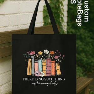 May include: Black tote bag featuring a colorful illustration of books with flowers. The text "THERE IS NO SUCH THING as too many books" is printed below the books. The bag has black handles and is displayed on a wooden bench.