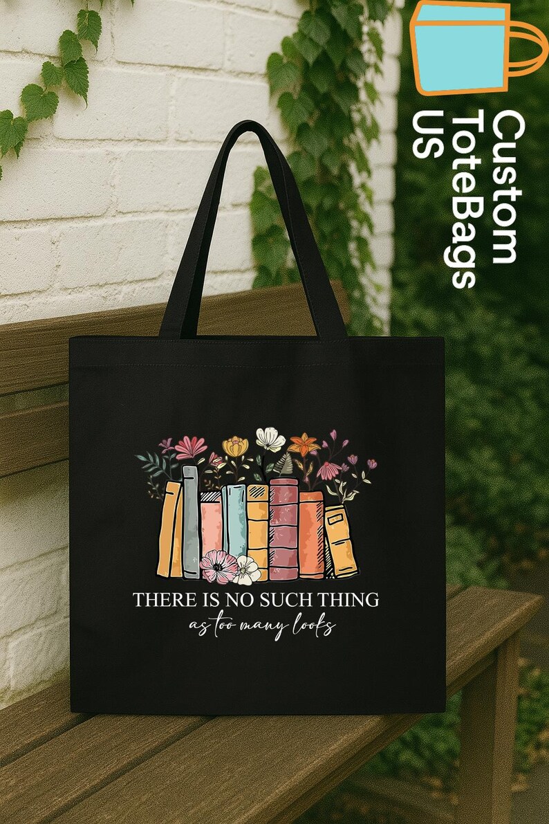May include: Black tote bag featuring a colorful illustration of books with flowers. The text "THERE IS NO SUCH THING as too many books" is printed below the books. The bag has black handles and is displayed on a wooden bench.