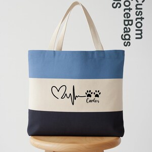 May include: A custom tote bag with a three-tone design: light blue, off-white, and navy blue. The off-white section features a heart rate graphic, paw prints, and the name "Carlos". The bag has cream-colored handles and the text "Custom Tote Bags US".