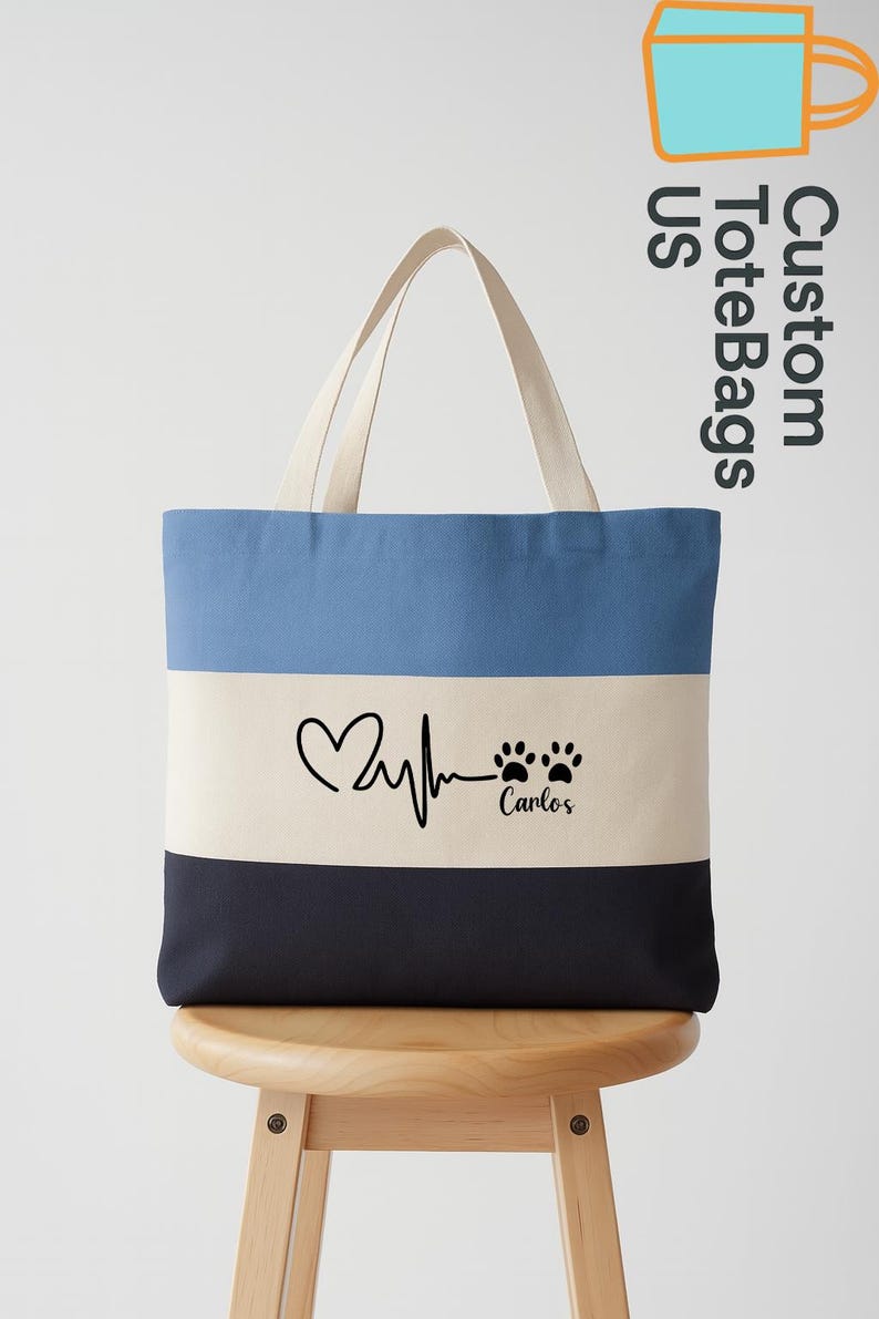 May include: A custom tote bag with a three-tone design: light blue, off-white, and navy blue. The off-white section features a heart rate graphic, paw prints, and the name "Carlos". The bag has cream-colored handles and the text "Custom Tote Bags US".