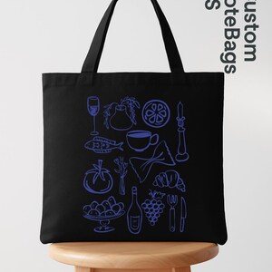 May include: Black tote bag with a blue line drawing of various food items, including a wine glass, fish, tomato, pasta, and grapes. The bag has a handle and the text "Custom Tote Bags US" in the upper right corner.