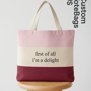 May include: A striped tote bag with a light pink, cream, and burgundy color scheme. The bag features the text "first of all i'm a delight" in black. The bag has cream-colored handles and is sitting on a wooden stool.