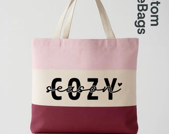May include: A multi-colored tote bag with the text "COZY season". The bag has a pink top, a cream-colored middle section, and a burgundy bottom. The bag is sitting on a wooden stool.