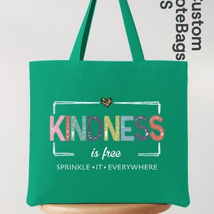 May include: A green tote bag with a white framed design that reads "KINDNESS is free. SPRINKLE IT EVERYWHERE." The word "KINDNESS" is in colorful letters. A small leopard print heart is above the word "KINDNESS".