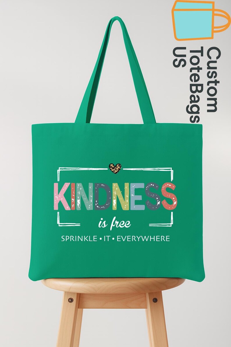 May include: A green tote bag with a white framed design that reads "KINDNESS is free. SPRINKLE IT EVERYWHERE." The word "KINDNESS" is in colorful letters. A small leopard print heart is above the word "KINDNESS".