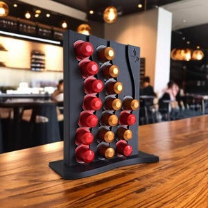 以下が含まれることがあります： 赤と金色のコーヒーポッドが入った、黒いコーヒーポッドホルダー。ホルダーは曲線的なデザインで、側面にコーヒーカップのアイコンが付いています。木製の表面に置かれています。背景にはぼやけたカフェの様子が写っています。
