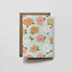 May include: A rectangular greeting card featuring a repeating floral pattern of peach, pink, and white flowers with green leaves on a light blue background, partially tucked into a plain brown kraft paper envelope.