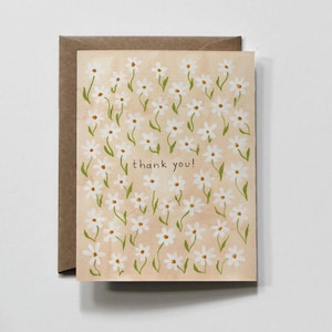 May include: A rectangular greeting card with a light tan background featuring a repeating pattern of white daisies with yellow centers and green stems. The text thank you! is printed in small black lowercase letters in the center of the card.