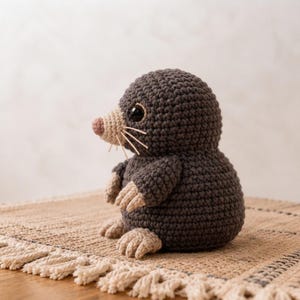 May include: A dark gray crocheted mole plush toy with a rounded body, a light beige snout, matching beige paws, and small black plastic eyes. The creature features thin, light-colored yarn whiskers extending from the sides of its nose.