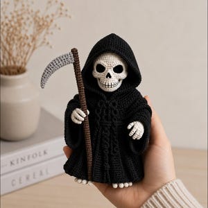 May include: A crocheted grim reaper figurine featuring a white skull head and hands, wearing a black hooded robe with a tied waist. The figure holds a brown staff topped with a grey curved scythe blade. The construction is detailed with fine yarn stitches throughout the entire piece, showcasing a dark aesthetic with contrasting light-colored skeletal features.